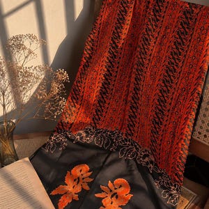 Puede incluir: Un tejido rectangular que presenta una sección con un vibrante patrón naranja y negro de rayas verticales y motivos complejos, que transiciona hacia un borde negro sólido adornado con dos grandes diseños florales bordados en color naranja.