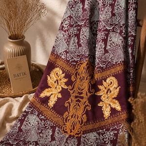 Op de afbeelding: Een diepbordeauxrode stof met batikprint, witte geometrische driehoekspatronen en complexe gouden bloemmotieven. Het textiel toont een centrale band met sierlijke, wervelende gouden ontwerpen tegen de donkerrode achtergrond.
