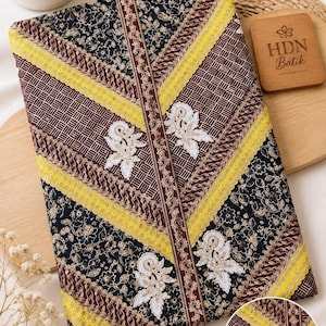 Könnte beinhalten: Ein gefaltetes Stoffstück mit einem dunkelbraunen und schwarzen Batikmuster, floralen Motiven, geometrischen Gitterabschnitten und vertikalen braunen Streifen. Das Textil ist mit leuchtend gelber Spitzenborte und weißer Blumenstickerei verziert.