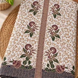 May include: A folded piece of white fabric featuring a brown floral pattern, dark purple and green botanical motifs, and a vertical geometric border. The bottom edge displays a dark blue grid pattern, and a wooden tag reads Batik Indonesia.