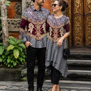 Puede incluir: Una camisa de manga larga con botones y una túnica a juego, ambas con un patrón inspirado en el batik en granate y gris con bordados florales dorados. La camisa se combina con pantalones negros, mientras que la túnica se lleva con pantalones negros.