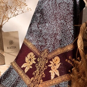 Puede incluir: Una pieza de tela doblada con un patrón batik, fondo color burdeos intenso, secciones geométricas azul claro y motivos florales dorados intrincados. Se ve una tarjeta rectangular con el texto BATIK PREMIUM QUALITY impreso.