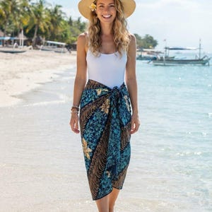 Puede incluir: Una camiseta sin mangas blanca combinada con un pareo largo con un estampado floral batik en azul oscuro y marrón. El pareo está atado a la cintura, creando una silueta drapeada. Se lleva un sombrero de paja de ala ancha con cinta negra.