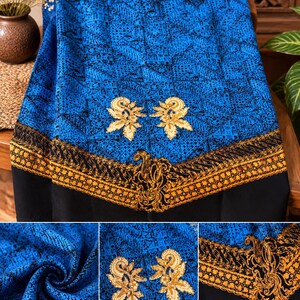 May include: A blue fabric featuring a black geometric grid pattern, accented by two gold floral appliques and a wide, ornate gold and black border along the bottom edge. The textile displays a rich, textured appearance with intricate detailing.