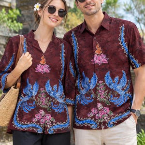 Puede incluir: Un conjunto a juego de camisas de batik color granate oscuro con un fénix azul vibrante, flores de loto rosas y motivos de llamas. Las camisas tienen cierre de botones, mangas cortas y un patrón detallado en toda la tela de estilo artesanal.