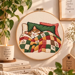 May include: A circular wooden embroidery hoop containing a cross-stitch pattern of a tabby cat sleeping under a colorful patchwork quilt. The quilt features a grid of green, red, and yellow squares with small floral motifs, and the cat rests on a green pillow.