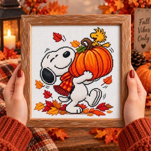 May include: A framed cross-stitch embroidery featuring the cartoon character Snoopy wearing a red and orange plaid scarf while carrying a large orange pumpkin, with a yellow bird sitting on top, surrounded by falling red and yellow autumn leaves.
