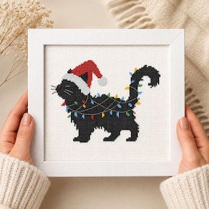 Puede incluir: Un marco cuadrado blanco que contiene un bordado de punto de cruz de un gato negro con un gorro de Papá Noel rojo y blanco. El gato está enredado en una cadena de luces navideñas de colores, con bombillas rojas, amarillas, azules y verdes.