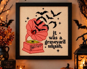 Puede incluir: Un bordado en punto de cruz enmarcado sobre tela beige que presenta un fonógrafo vintage rosa con un disco verde, rodeado por siluetas negras de murciélagos volando y pequeñas estrellas, con el texto "it was a graveyard smash" en negro.
