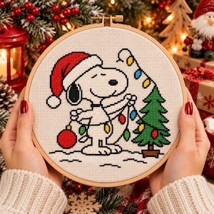 May include: A circular wooden embroidery hoop containing a cross-stitch pattern of a white dog wearing a red Santa hat. The dog is decorating a green Christmas tree with colourful string lights and holding a large red ornament on a white background.