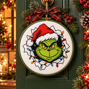 May include: A circular embroidery hoop containing a cross-stitch pattern of a green character wearing a red Santa hat, surrounded by colorful Christmas lights. The design is framed in a dark green hoop and topped with a plaid fabric bow.