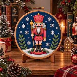 Può includere: Un telaio da ricamo circolare in legno contenente un disegno a punto croce di un soldato schiaccianoci in uniforme rossa che tiene in mano un bastone dorato. La figura si trova su una base bianca innevata con foglie verdi e bacche rosse.