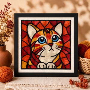 Può includere: Un ricamo a punto croce quadrato raffigurante un gatto tigrato rosso con grandi occhi blu, su uno sfondo geometrico di forme rosse e arancioni in stile vetrata, il tutto racchiuso in una semplice cornice nera da appendere.