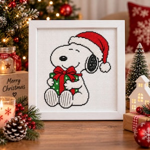 May include: A square white frame containing a cross-stitch pattern of a white cartoon dog with a black ear, wearing a red and white Santa hat, and holding a green gift box with a red ribbon and bow. The design is on a white grid fabric.