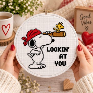 Puede incluir: Un bastidor de bordado circular con un diseño de punto de cruz de un perro blanco con un pañuelo rojo mirando a través de un telescopio marrón, con un pequeño pájaro amarillo encima. El texto negro dice LOOKIN' AT YOU.