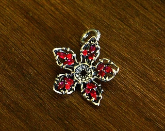 Peut inclure: Un pendentif en métal argenté en forme de fleur à cinq pétales. Chaque pétale est orné de plusieurs petites pierres rouges taille marquise. Le centre de la fleur présente un groupe de minuscules pierres claires et une bélière circulaire.