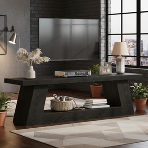 May include: A long, black wooden media console featuring a thick top surface and a lower shelf. The console has angled, sturdy legs and a visible wood grain texture. It is positioned horizontally, providing a flat surface for electronics and decor.