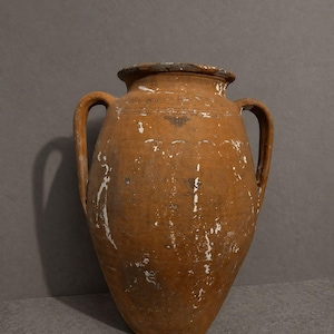May include: A large, terracotta-colored earthenware vessel featuring two side handles, a wide circular rim, and a tapered base. The surface displays a weathered texture with visible patches of white paint residue and subtle horizontal incised lines.