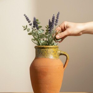 May include: A terracotta pitcher with a textured, matte orange body and a glazed, speckled olive-green rim. The pitcher features a single side handle and contains a bouquet of purple lavender sprigs and green eucalyptus leaves.