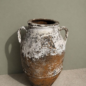 May include: A large, weathered terracotta vessel with two side handles. The exterior features a textured, distressed finish with a combination of deep brown clay tones and patches of peeling white paint, creating a mottled, aged appearance.
