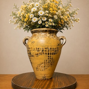 May include: A yellow ceramic vase with two handles and a dark grid pattern, filled with a bouquet of white and yellow daisies. The vase features a distressed, textured finish with visible brushstrokes and dark accents around the rim and base.