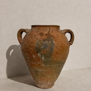 May include: An antique terracotta amphora with a weathered, textured surface in shades of brown and grey. The vessel features two side handles, a wide circular opening, and a tapered base, with a visible vertical crack extending from the rim.