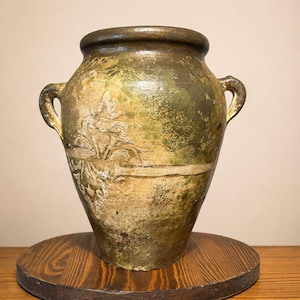 May include: A large, weathered ceramic urn featuring a distressed finish in shades of olive green, brown, and cream. The vessel has two side handles, a wide rim, and a raised, embossed floral relief pattern on the front surface of the body.