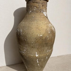 May include: A tall, weathered earthenware vessel with a tapered base and a wide, rounded body. The surface is a muted, earthy tan colour with visible pitting, texture, and patches of white residue throughout the exterior, featuring a thick rim.