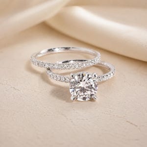 Op de afbeelding: Een zilverkleurige verlovingsringset met een grote, rondgeslepen centrale steen die door vier pootjes wordt vastgehouden, en een bijpassende pavé-ring. Beide ringen zijn bezet met kleine, heldere en fonkelende steentjes.