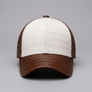 Peut inclure: Une casquette de baseball avec un panneau avant blanc et des panneaux latéraux marron, tous dotés d'une texture en peau de crocodile. La visière incurvée est également marron avec la même texture, et un petit bouton est au sommet.
