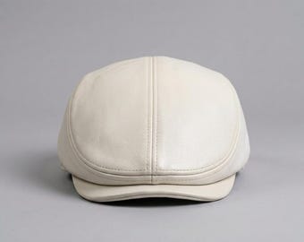 May include: A cream-colored flat cap constructed from smooth leather. The cap features a rounded crown with a central vertical seam running from the top to the front brim, showcasing a clean, structured design with a short, curved visor.