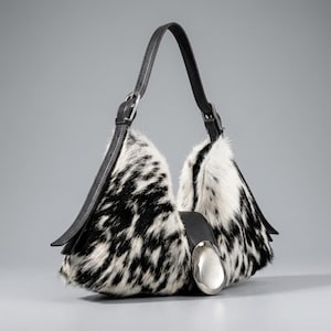 May include: A hobo-style handbag featuring a black and white cowhide pattern with fur texture. The bag includes a black leather shoulder strap with an adjustable silver-tone buckle and a large, smooth, oval-shaped silver-tone metal accent on the front.