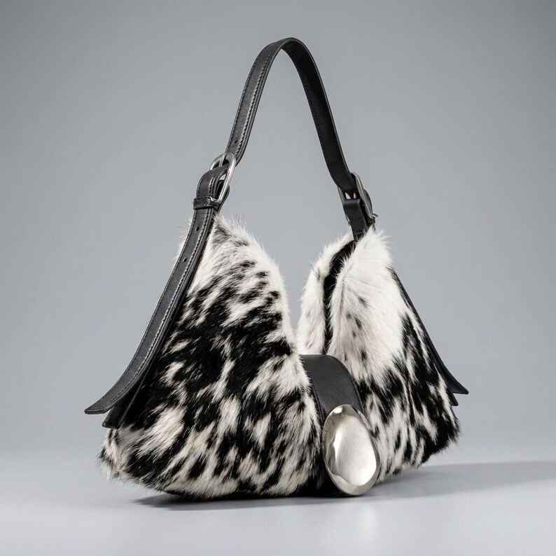 May include: A hobo-style handbag featuring a black and white cowhide pattern with fur texture. The bag includes a black leather shoulder strap with an adjustable silver-tone buckle and a large, smooth, oval-shaped silver-tone metal accent on the front.