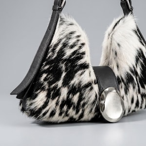 May include: A shoulder bag featuring a black and white cow print faux fur texture. The bag has a smooth black leather strap and a large, circular, polished silver-tone metal accent piece centered on the front flap closure mechanism.