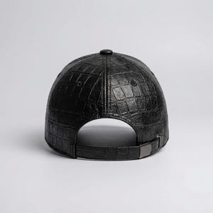 Peut inclure: Une casquette de baseball noire avec un motif texturé en peau de crocodile sur toute la surface. La casquette comprend un bouton noir assorti au centre supérieur et une sangle réglable à l'arrière avec une boucle métallique foncée.