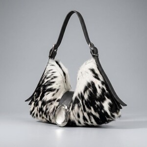 May include: A hobo-style handbag featuring a black and white cowhide pattern with fur texture. The bag has a single black leather strap, black leather side panels, and a large, smooth, oval-shaped silver-toned metal accent centered on the front.