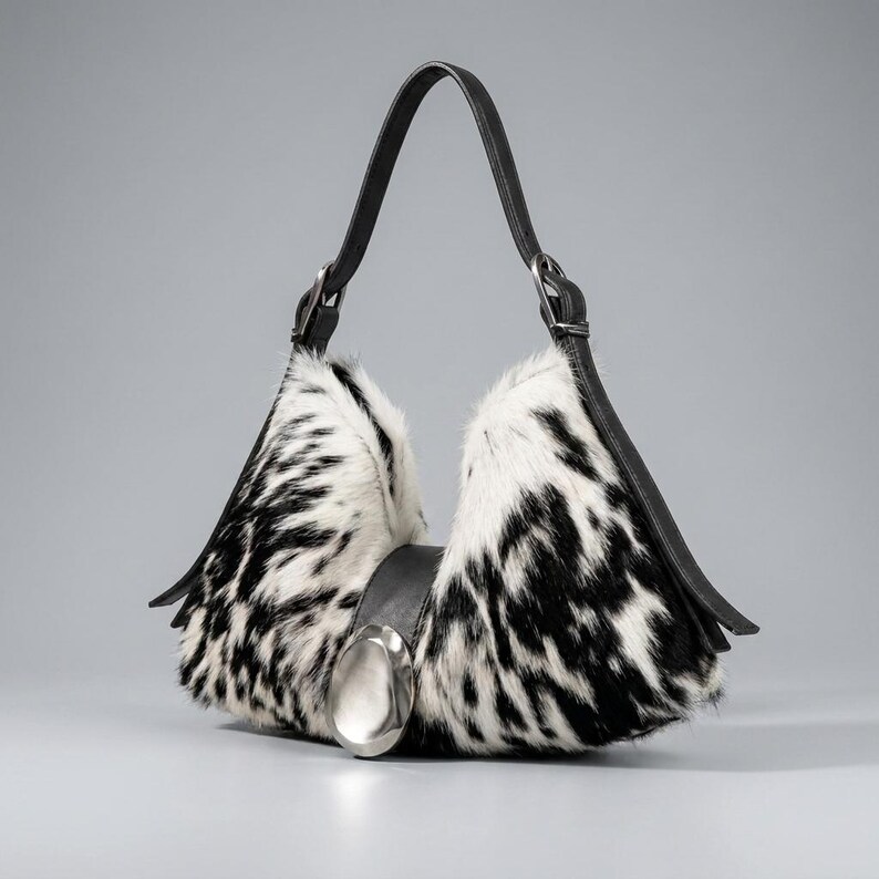May include: A hobo-style handbag featuring a black and white cowhide pattern with fur texture. The bag has a single black leather strap, black leather side panels, and a large, smooth, oval-shaped silver-toned metal accent centered on the front.