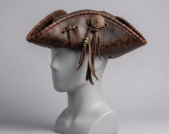 May include: A brown tricorn hat with a weathered texture, featuring a decorative brass-colored compass emblem, leather cord lacing, and a small skull charm dangling from the side. The brim is stitched along the edges with brown cord.