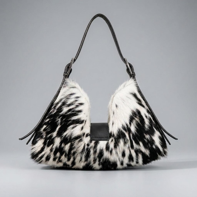 May include: A crescent-shaped shoulder bag featuring a black and white cow-print fur texture. The bag includes a smooth black leather strap attached with silver-toned hardware and a central black leather tab closure on the front exterior.