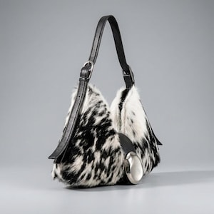 May include: A hobo-style handbag featuring a black and white cowhide-patterned fur exterior. The bag includes a single black leather shoulder strap with an adjustable silver-tone buckle and a large, smooth, polished silver-tone metal accent piece.