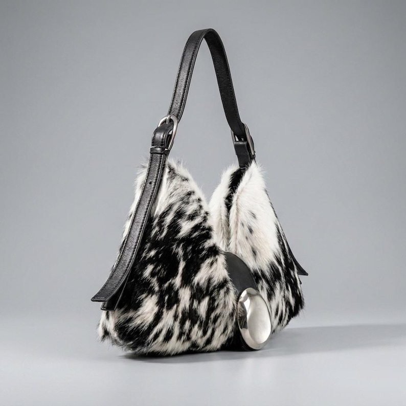 May include: A hobo-style handbag featuring a black and white cowhide-patterned fur exterior. The bag includes a single black leather shoulder strap with an adjustable silver-tone buckle and a large, smooth, polished silver-tone metal accent piece.