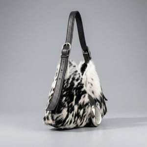 May include: A shoulder bag featuring a black and white spotted animal print fur texture. The bag has a black leather strap with a silver-toned metal buckle for adjustment and a large, smooth, silver-toned metal accent piece on the side.