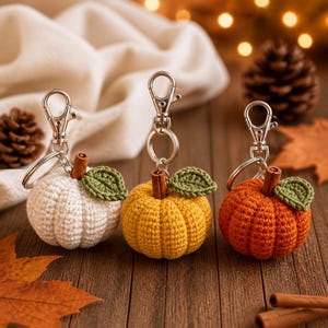 May include: Three crochet pumpkin keychains in white, yellow, and orange. Each pumpkin features a small brown stem, a single green crocheted leaf, and a silver-toned metal lobster clasp attached to a circular ring for secure fastening.