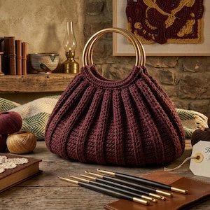 May include: A deep burgundy hand-crocheted pouch with a pleated texture and two circular gold-toned metal handles. A small white tag attached to the pouch features the text: The Aura Pouch - Hand-Crocheted, Artisanal. 380 is printed below the text.