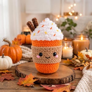 Peut inclure: Un jouet en peluche crocheté en forme de tasse à café avec une base orange vif, une manchette beige et une garniture fouettée blanche. La tasse présente un visage souriant avec des yeux noirs, des joues roses et deux bâtonnets marron.