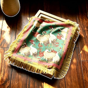 May include: A square fabric textile featuring a teal green background with a repeating pattern of white cranes and red floral accents. The textile is bordered by a decorative red and gold frame and finished with long yellow fringe on all four sides.
