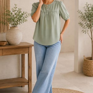 Pode incluir: Uma blusa verde-sálvia de manga curta com decote redondo, carcela de botões parcial e mangas bufantes franzidas, combinada com calças de ganga largas azul-claras e sapatos rasos beges com um pequeno laço decorativo na parte frontal.