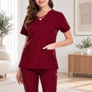 May include: A solid burgundy scrub top featuring short sleeves, a V-neckline with a decorative crisscross strap detail, and two large patch pockets on the front. The matching burgundy scrub pants feature side pockets and a straight-leg design.