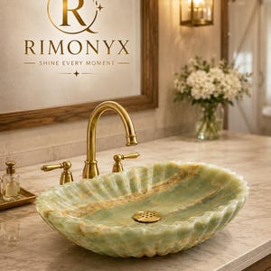May include: An oval, scalloped-edge vessel sink crafted from light green and tan natural stone with a polished finish. It features a central gold-toned drain and is paired with a matching gold-toned arched faucet and two side handles.