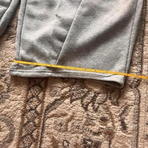 Peut inclure: Un pantalon de jogging gris clair avec une coupe droite et un ourlet cousu. Un m&egrave;tre ruban jaune est plac&eacute; horizontalement sur le bas de la jambe, indiquant une largeur d'environ 26,7 centim&egrave;tres &agrave; cet endroit pr&eacute;cis du v&ecirc;tement.